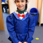 Girl in a blue dress with white ruffled collar and red bow tie, wearing a small black hat with a blue ribbon and flower; standing in a classroom.
