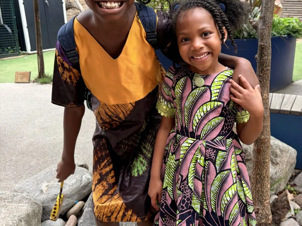 Two young girls pose together outdoors, smiling, wearing colorful patterned dresses and black shoes with white socks on a rocky garden path.
