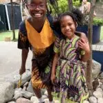 Two young girls pose together outdoors, smiling, wearing colorful patterned dresses and black shoes with white socks on a rocky garden path.